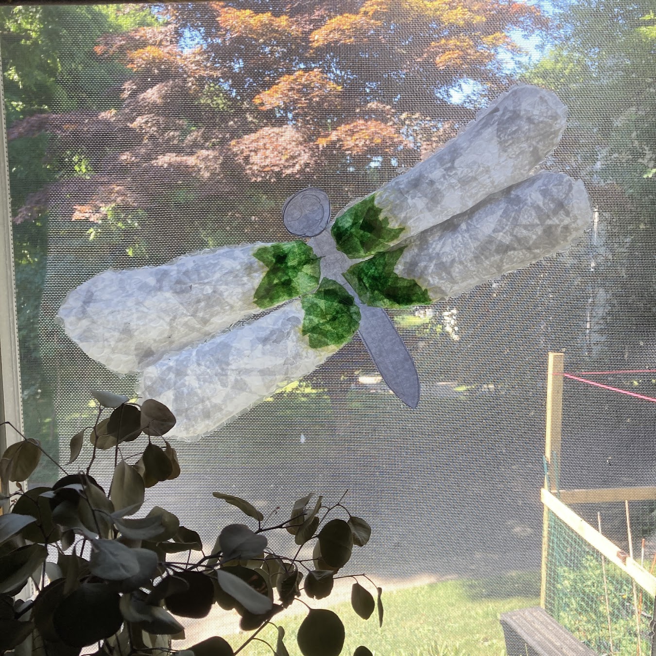 Completed stained glass dragonfly hanging in a window with a eucalyptus plant in the foreground.